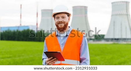 Nuclear power plant engineer with a tablet. A man with a beard in a protective helmet and an orange vest stands against the background of a nuclear power plant. Work inspection Royalty-Free Stock Photo #2312931033