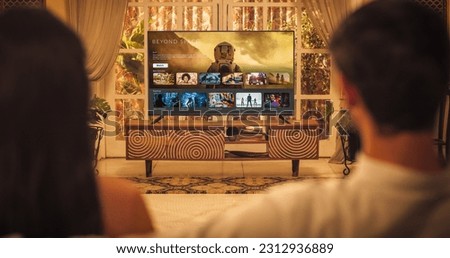 Back View of Young Indian Couple Watching TV while Sitting on a Couch in the Living Room. Relaxed Evening for Enjoying Favourite Movies, TV Shows, Content and Ads Thank to Streaming Online Service