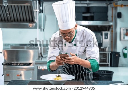 Asian male chef cooks macaroni and decorates the dish beautifully with vegetables to look appetizing before serving to customers using smartphones to take pictures of the food in the restaurant
