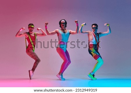 Sport show. Three funny men in colorful sportswear doing aerobics exercises against gradient blue pink studio background in neon light. Concept of sportive and active lifestyle, humor, retro style. Ad Royalty-Free Stock Photo #2312949319