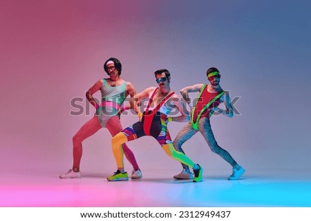 Funny men in vintage, colorful sportswear posing, doing aerobics exercises against gradient blue pink studio background in neon light. Concept of sportive and active lifestyle, humor, retro style. Ad Royalty-Free Stock Photo #2312949437