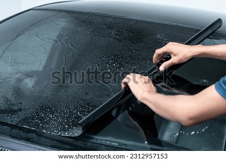 Male auto specialist worker hand rolling car window film on front windscreen glass surface. Car side window film removal and tinting installation.  Royalty-Free Stock Photo #2312957153