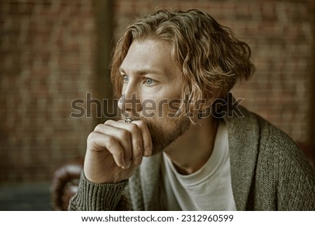 Portrait of a handsome forty-year-old man with curly hair, dressed in a casual knitted cardigan, who looks thoughtfully to the side. Loft interior. People, emotions. Life style. Royalty-Free Stock Photo #2312960599