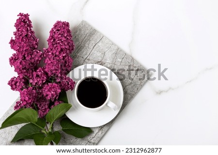Branch of burgundy lilac flowers and cup of fresh black coffee on white background with a marble texture with a place for text. Flat lay, copy space