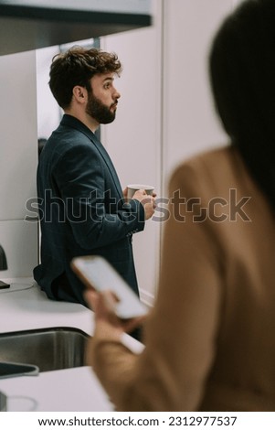 A side view photo of handsome male person talking to his colleague while holding a cup of tea. His female coworker is checking her phone