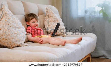 The joyful child is seated on the sofa holding the TV remote, watching cartoons with a beaming smile. A little baby is lying on the couch, watching a cartoon on the television set with happiness.
