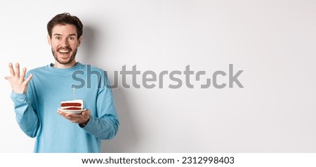 Excited man receive birthday surprise, holding bday cake and smiling happy, standing over white background, making wish on lit candle.