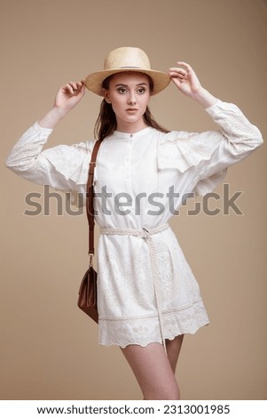 High fashion photo of beautiful elegant young woman in a pretty white dress with the sleeves, hat, handbag posing over beige brown background. Studio Shot, portrait.