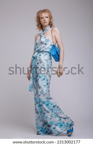 High fashion photo of beautiful elegant young woman in a pretty rich blue jumpsuit with floral pattern, handbag, clutch posing over white, soft gray background. Studio Shot, portrait. Blonde