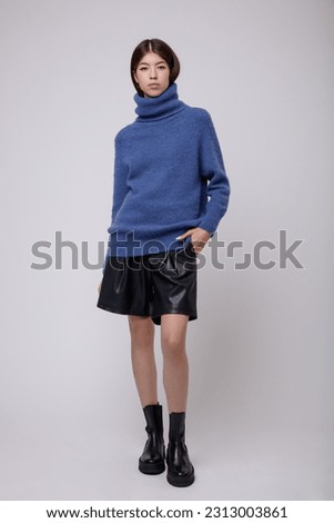 Fashion photo of a beautiful elegant young asian woman in a pretty blue sweater, blouse, black leather shorts posing on white, soft gray background. Studio Shot, portrait. Slim figure. 