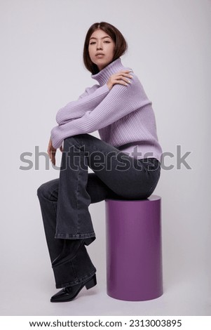Fashion photo of a beautiful elegant young asian woman in a pretty pale purple, lilac, lavender sweater, blouse, black jeans, denim posing on white, soft gray background. Model sits  on cube, cylinder