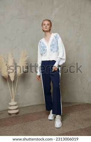 Fashion photo of a beautiful elegant young woman in a pretty white sweater, sport pants, trousers with the stripes posing over sandy beige textured wall. Studio Shot. Blonde