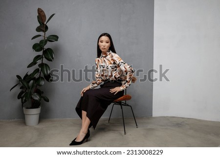 High fashion photo of beautiful elegant young asian woman in pretty beige shirt with pattern, long black skirt posing on textured gray wall, tall flower. Slim figure. Model is sitting on a chair.  Royalty-Free Stock Photo #2313008229