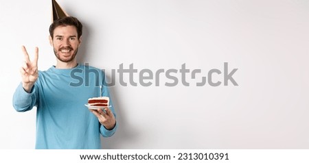 Celebration and holidays concept. Happy young man celebrating birthday, taking picture with peace sign, wearing party hat and holding bday cake, white background.