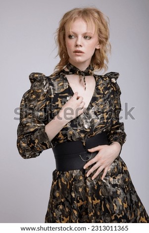 High fashion photo of beautiful elegant young woman in a pretty military camouflage dress with sleeves, boots, wide belt posing over white, soft gray background. Studio Shot, portrait. Blonde