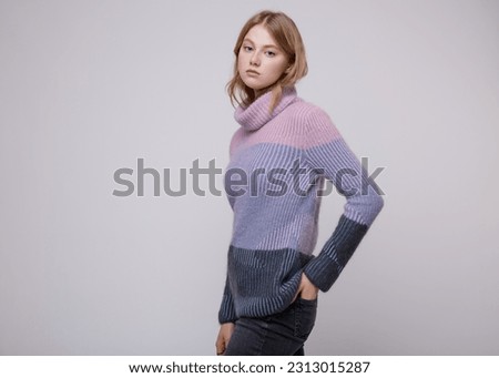 Fashion photo of beautiful elegant young woman in a pretty triple color pink, blue, gray sweater, blouse, black jeans, denim posing on white, soft gray background. Blonde