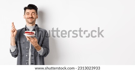 Hopeful birthday guy making wish with fingers crossed, holding bday cake on party, standing against white background.