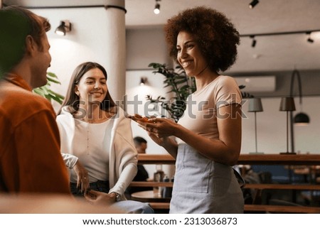 Beautiful african waitress wearing apron smiling while taking order in cafe Royalty-Free Stock Photo #2313036873
