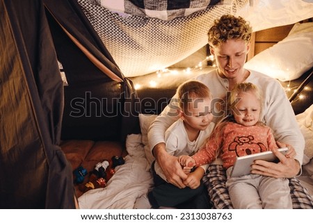 Smiling dad and his cute kids wearing pajamas watching cartoons on digital tablet while sitting in handmade tent in children's room