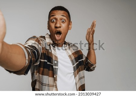 Young brunette man in shirt screaming and gesturing at camera while taking selfie photo isolated over white background