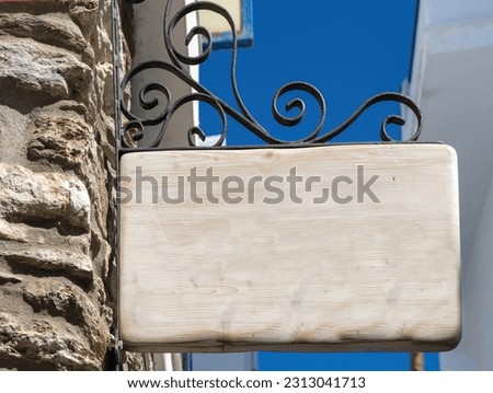 Empty wooden sign hanging from a metal structure. Signboard on stonewall blue sky Cyclades island Greece. Close up under view copy space ad template