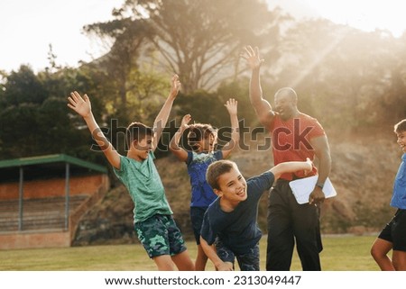 Elementary school kids celebrating with their sports coach during PE. Male trainer motivating a group of primary school kids. Sports and recreation in school. Royalty-Free Stock Photo #2313049447