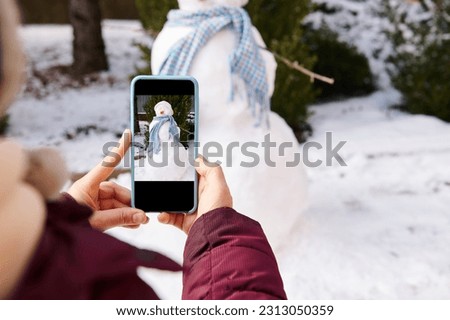 Close-up of hands holding smart mobile phone in live view mode, photographing snowman with blue shawl and carrot nose, in a snow covered nature background. Wintertime. Christmas time. Winter leisures