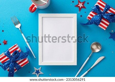 Embrace spirit of Independence Day at this festive gathering. Top view of table arrangement featuring cutlery, cup, stars, confetti, gift boxes. Blue backdrop with empty photo frame for ads or picture