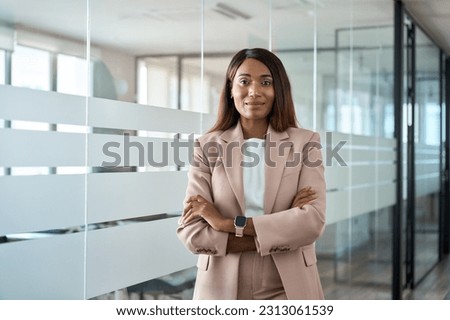 Confident proud young professional business woman ceo, female corporate executive leader, African American lady lawyer wearing suit standing arms crossed in office near glass wall, portrait.