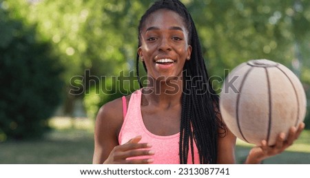Portrait of Strong Black Young Woman Tossing a Basket Ball While Looking at the Camera in an Outdoor Court. Smiling Female Athlete Defying Stereotypes and Following her Dream of Going Professional Royalty-Free Stock Photo #2313087741