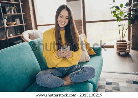 Full length photo of gorgeous positive lady sit couch crossed legs use smart phone chatting communicate modern flat inside