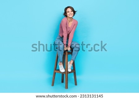 Full length photo of optimistic friendly girl wear pink cardigan denim trousers sitting on chair smiling isolated on blue color background