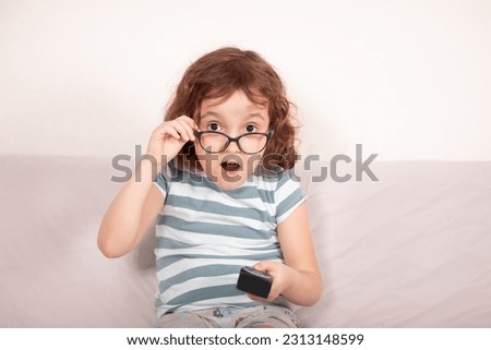 Cute curly little girl kid child in a eyeglasses surprised watching tv at home with remote control in a hands