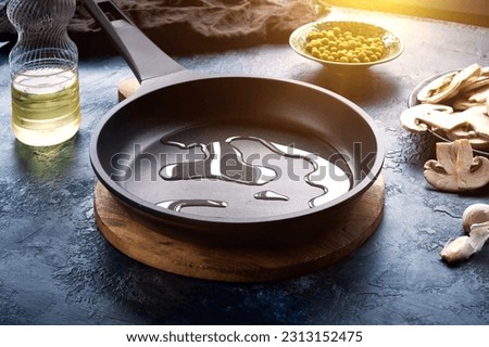 Dark frying pan with poured oil on a dark blue abstract background. Nearby is a plate of champignons, green peas, and a bottle of oil. Template for design. Royalty-Free Stock Photo #2313152475