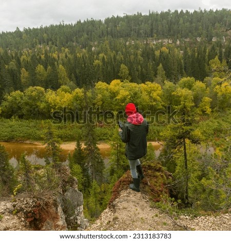 a woman in a jacket and hat and rubber boots stands on the edge of a cliff against the background of a forest and takes pictures of the landscape on her smartphone. the concept of travel and tourism