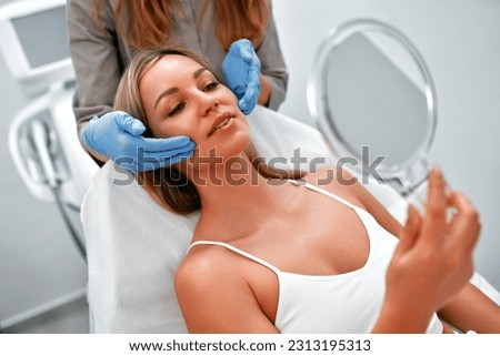 Cosmetologist is consulting woman client who looking at mirror in beauty clinic. Beauty industry concept. Beautician preparing patient to skin care procedure in spa salon Royalty-Free Stock Photo #2313195313