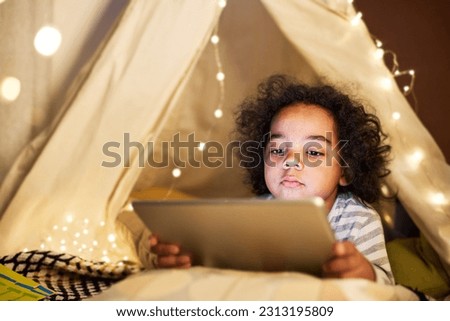 Cute child with curly hair watching cartoon on digital tablet while lying on the floor in handmade tent