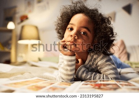 Portrait of cute child with curly hair looking at camera while lying on bed with book