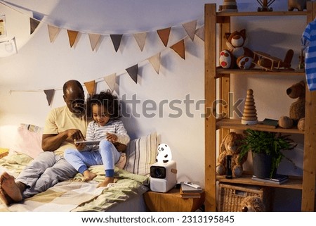 African American dad using digital tablet together with his little son before sleep in bedroom