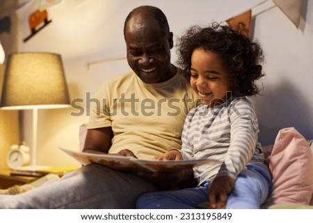 Happy dad showing pictures in book to his little son while reading him before sleep at night