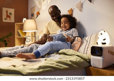 African American child watching funny cartoon on digital tablet together with his dad before sleep in the night