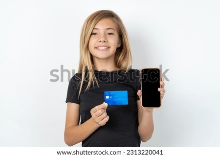 Photo of adorable beautiful caucasian teen girl wearing sportswear ball over white wall holding credit card and Smartphone. Reserved for online purchases