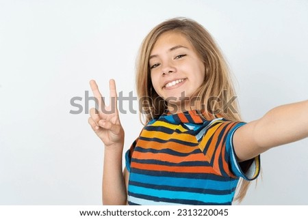 Positive beautiful caucasian teen girl wearing striped colorful T-shirt over white wall take selfie make v-sign
