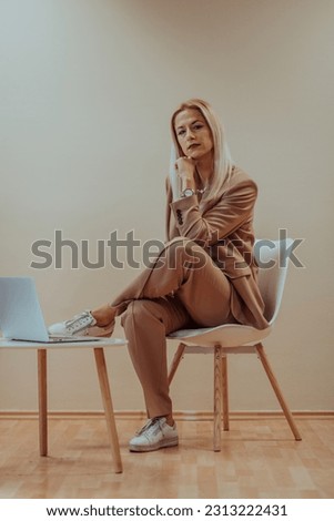 A professional businesswoman sits on a chair, surrounded by a serene beige background, diligently working on her laptop, showcasing dedication and focus in her pursuit of success