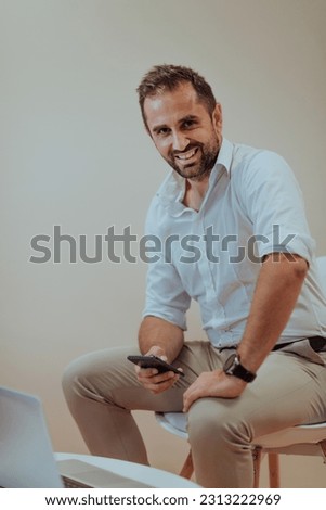 A confident businessman sitting and using laptop and smartphone with a determined expression, while a beige background enhances the professional atmosphere, showcasing his productivity and expertise.