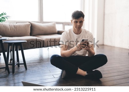 Young male sitting on floor and browsing mobile phone while looking at screen for text messages while passing free time in cozy living room at home