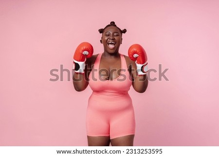 Portrait of emotional African American woman, professional boxer wearing red boxing gloves training, screaming  isolated on pink background. Sport, motivation, body positive concept