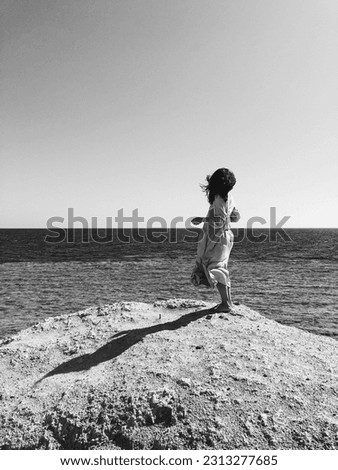 A black and white photo captures a girl standing on a rocky shore by the sea, her hair dancing in the wind