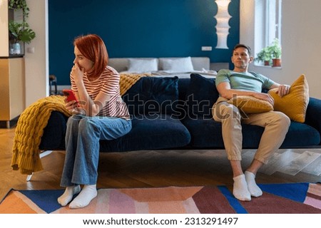 Conflict, quarrel. Haughty husband sit on couch, feeling annoyed, offended. Woman with smartphone feeling ignored, misunderstood. Jealous neurotic man tyrant, abuser, humiliates wife. End relationship Royalty-Free Stock Photo #2313291497