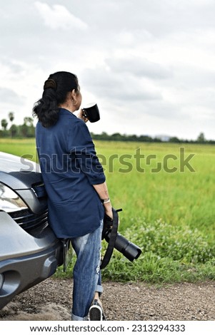 black hair woman photographer standing by her car hold a mug of coffee and her big camera